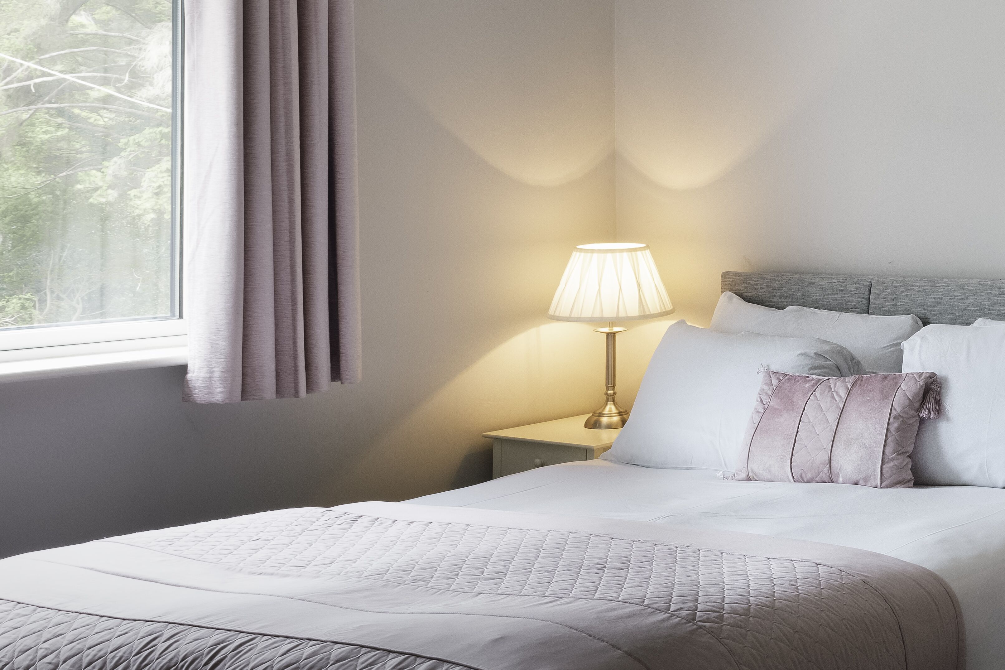 Una cama bien hecha con un edredón acolchado, una lámpara de noche y una ventana con cortinas.