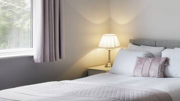 Una cama bien hecha con un edredón acolchado, una lámpara de noche y una ventana con cortinas.