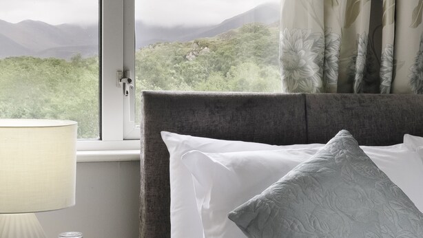 A bedroom with a bed, a nightstand with a lamp, and a view of mountains through a window. - The Climbers Inn (Glencar)