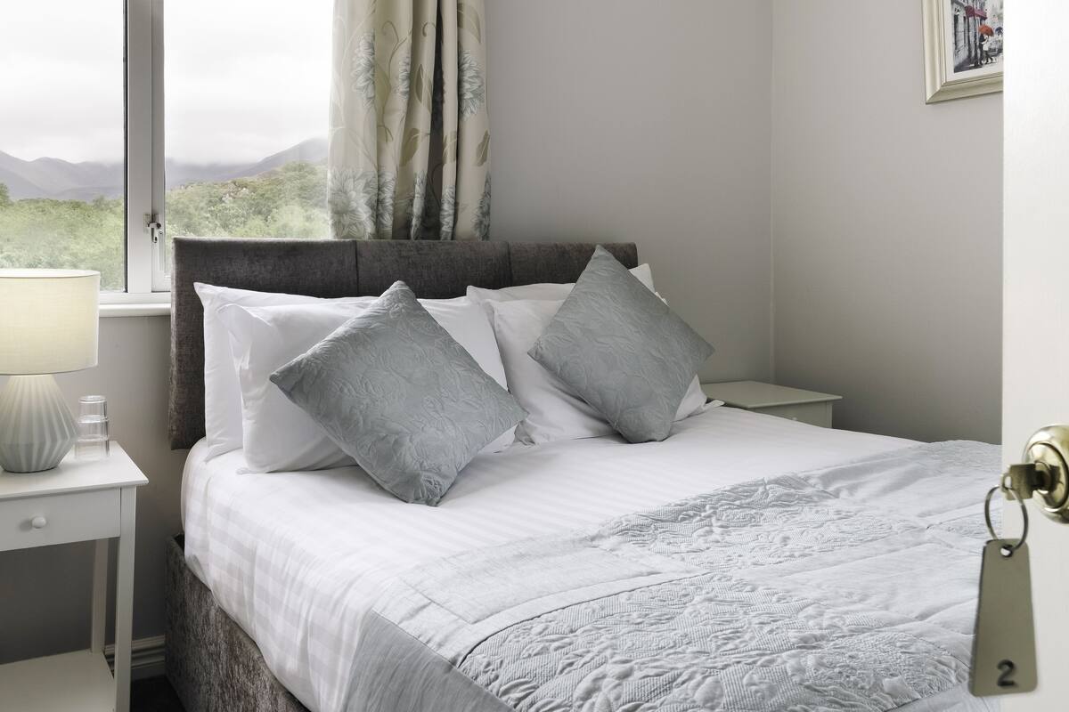 A neatly made bed with white and gray linens, a gray headboard, and a view of mountains through a window.