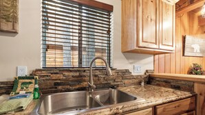 A kitchen with a granite countertop, stainless steel sink, wooden cabinets, and a window with blinds.