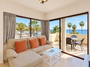 Ein modernes Wohnzimmer mit einem beigefarbenen Sofa, einem Glastisch und Blick auf das Meer und Palmen.