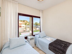 A hotel room with two beds, a nightstand, and a window with a view of the beach and palm trees.