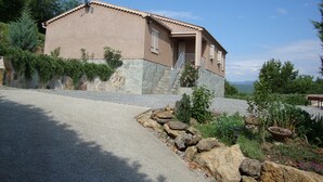 A two-story house with a stone foundation and a tiled roof, surrounded by a gravel driveway and a stone wall with vegetation. - Beautiful newly built house with private pool in the Ardèche (Chassagnes)