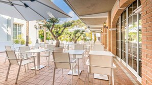 An outdoor patio with white tables and chairs, covered by large umbrellas, and surrounded by brick walls and windows. - Park Hotel Pineta (Caorle)