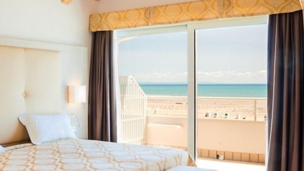 A bedroom with a bed, bench, and a large window offering a view of the beach. - Park Hotel Pineta (Caorle)