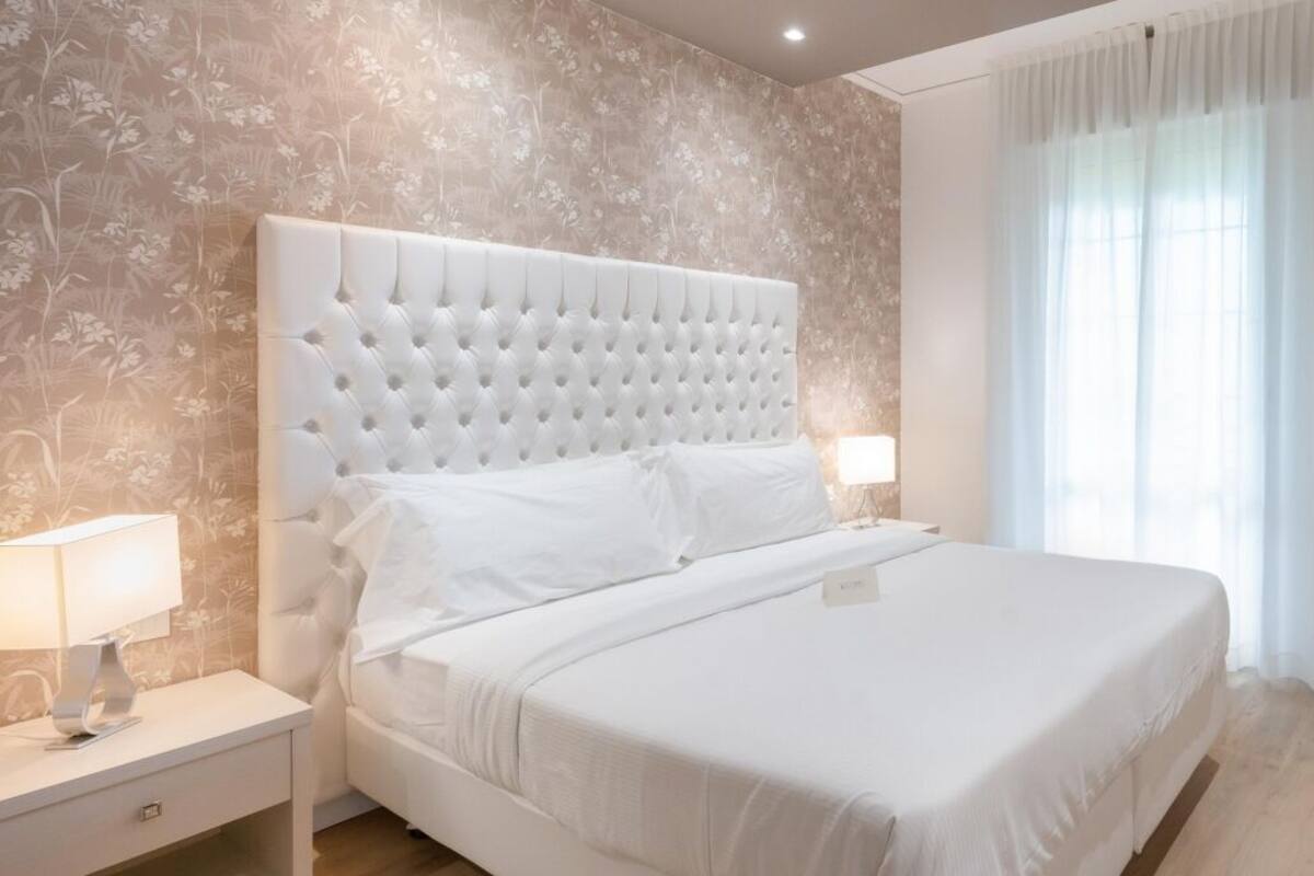 A modern bedroom with a tufted headboard, a large bed, bedside tables, and a window with sheer curtains.