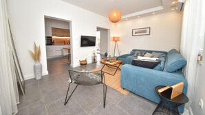 Una sala moderna con un sofá azul, una mesa de centro y una silla negra. Se ve una cocina al fondo, con un televisor montado en la pared.