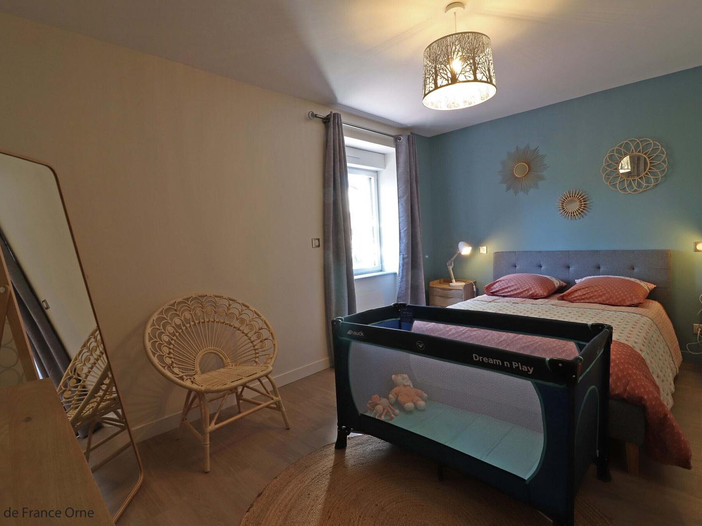 Une chambre à coucher avec un lit, un berceau, une chaise en osier et un luminaire fixé au plafond.