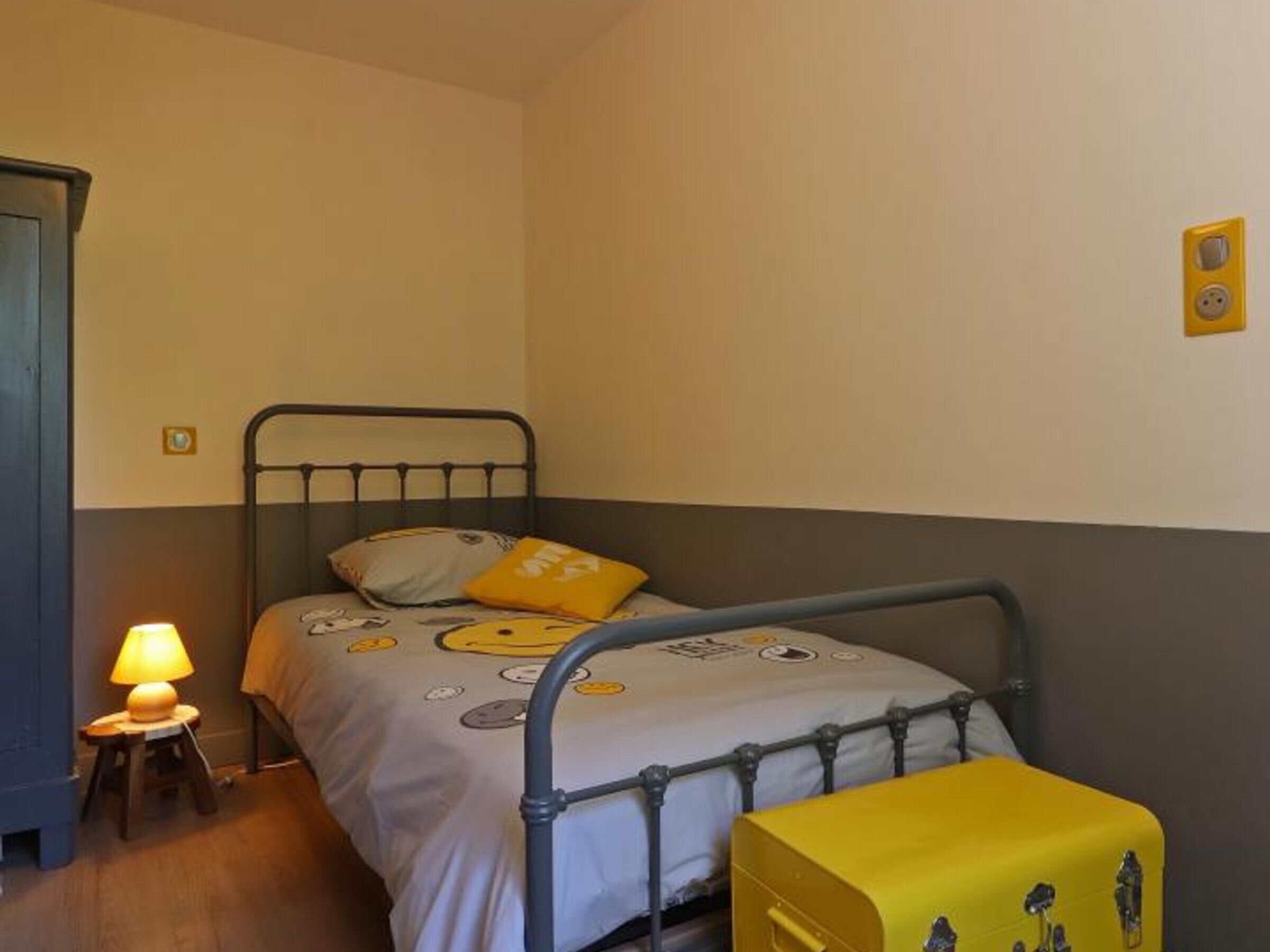 Een enkel bed met metalen frame, een nachtkastje met lamp en een gele opbergbox.
