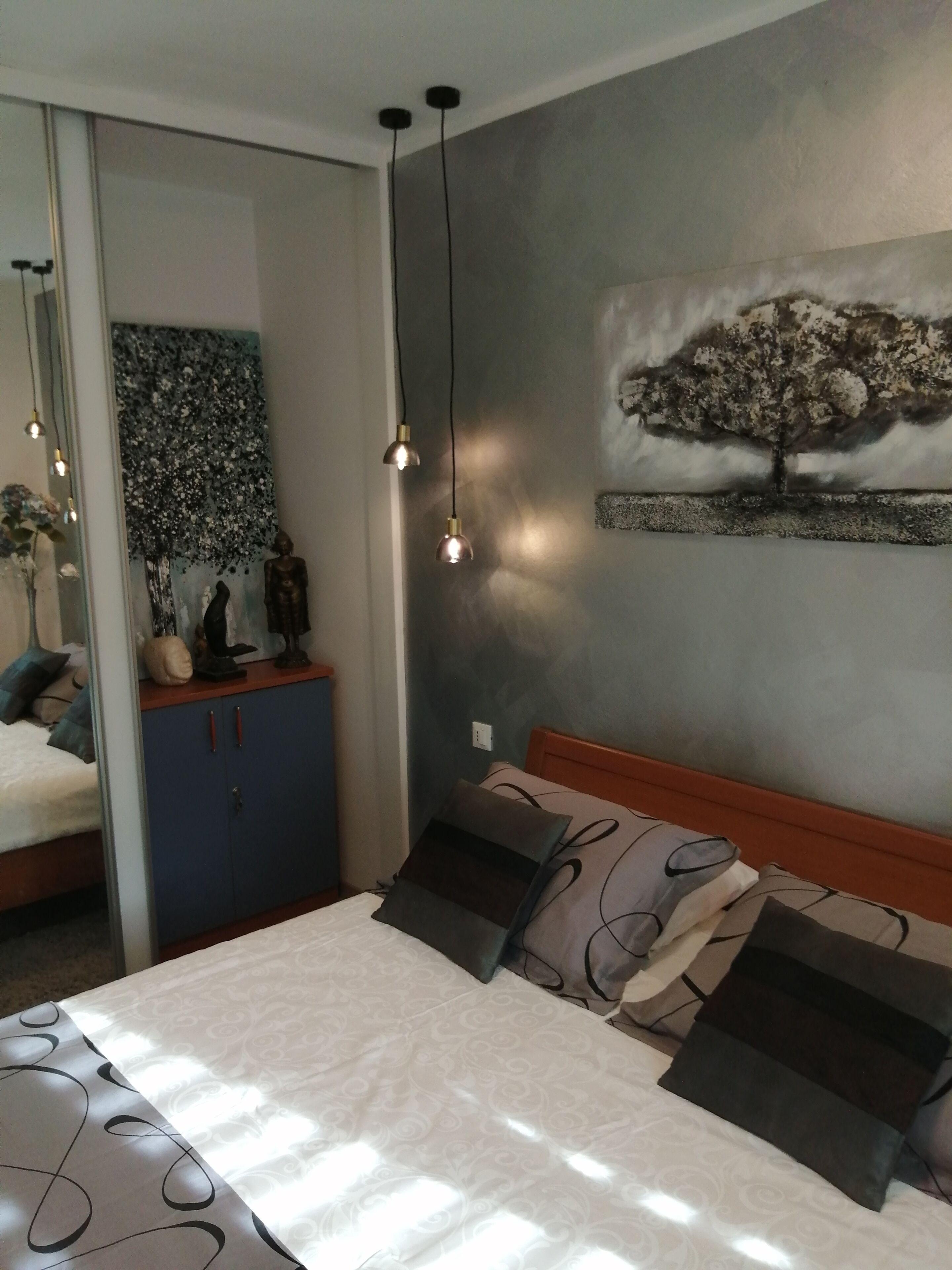 A bedroom with a bed, a dresser, a mirror, and a wall-mounted artwork of a tree.
