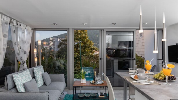 Una sala de estar moderna con un sofá gris, un comedor con mesa para dos, y una cocina con televisión.