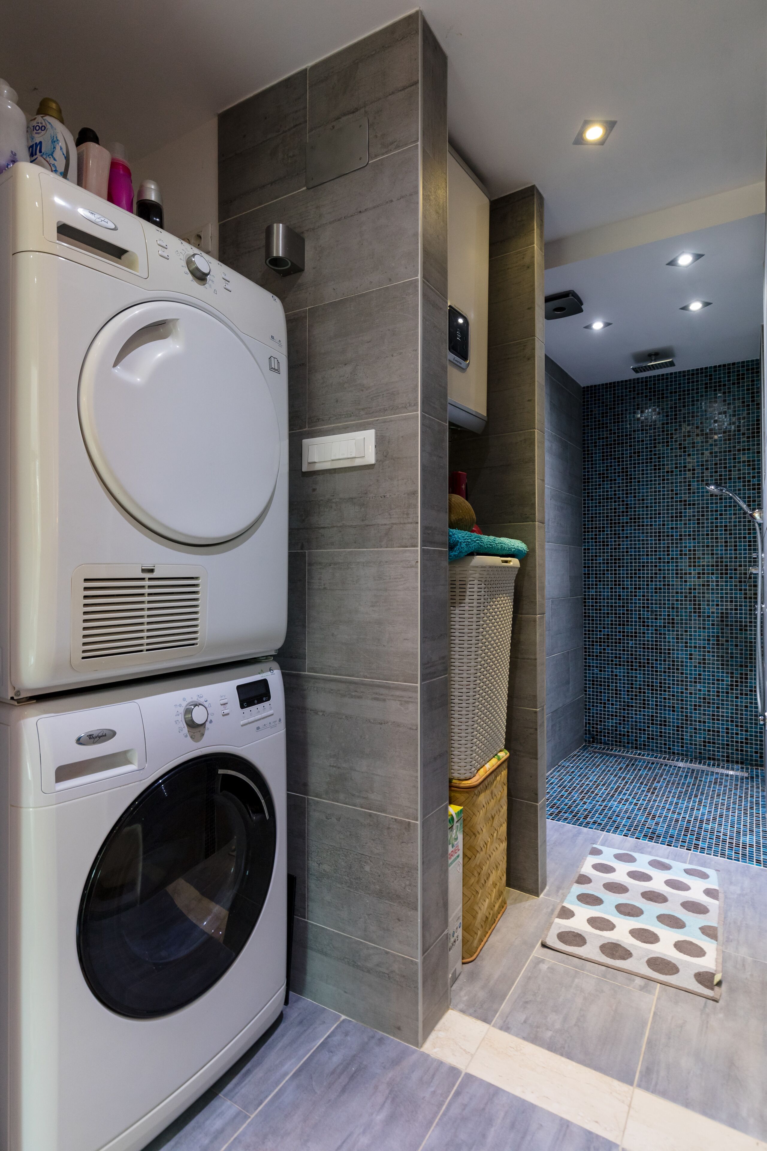 A modern bathroom with a stacked washer and dryer, a shower with blue mosaic tiles, and a small step leading to a basket.