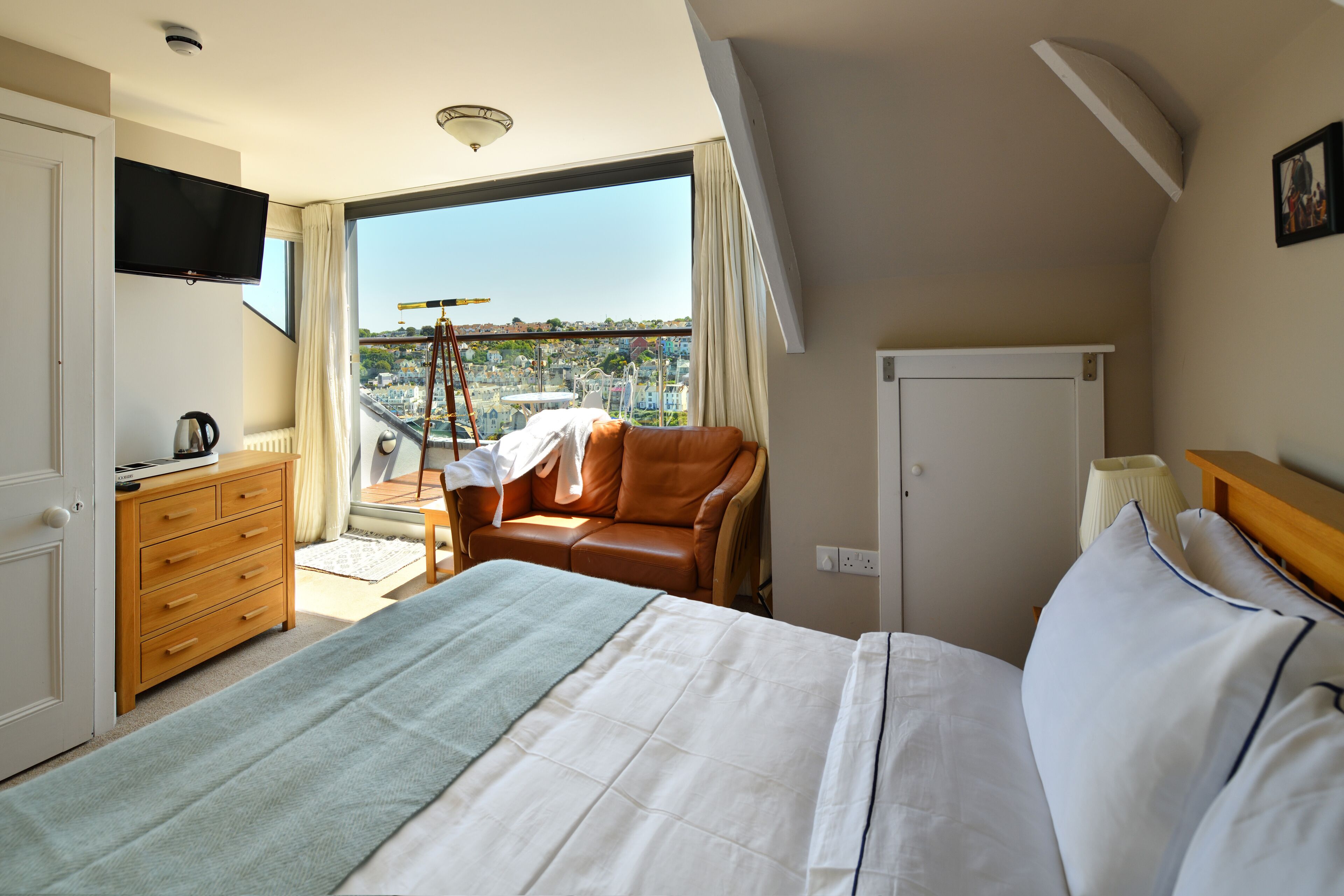 A hotel room with a large bed, a TV mounted on the wall, a view of the city through a large window, and a wooden dresser.