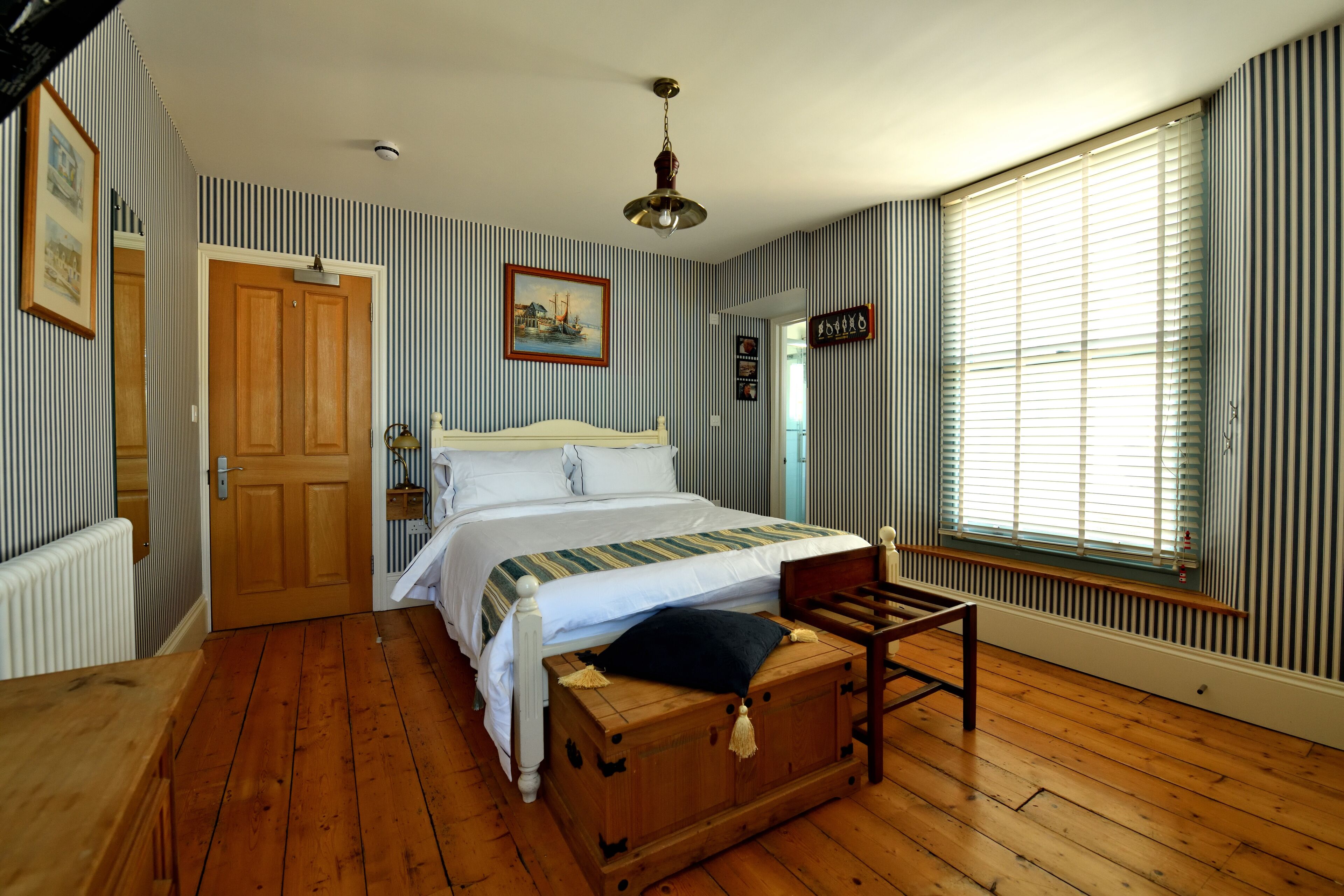 A bedroom with a wooden floor, a bed with a white headboard, a chest of drawers, a wooden bench, and a window with blinds.