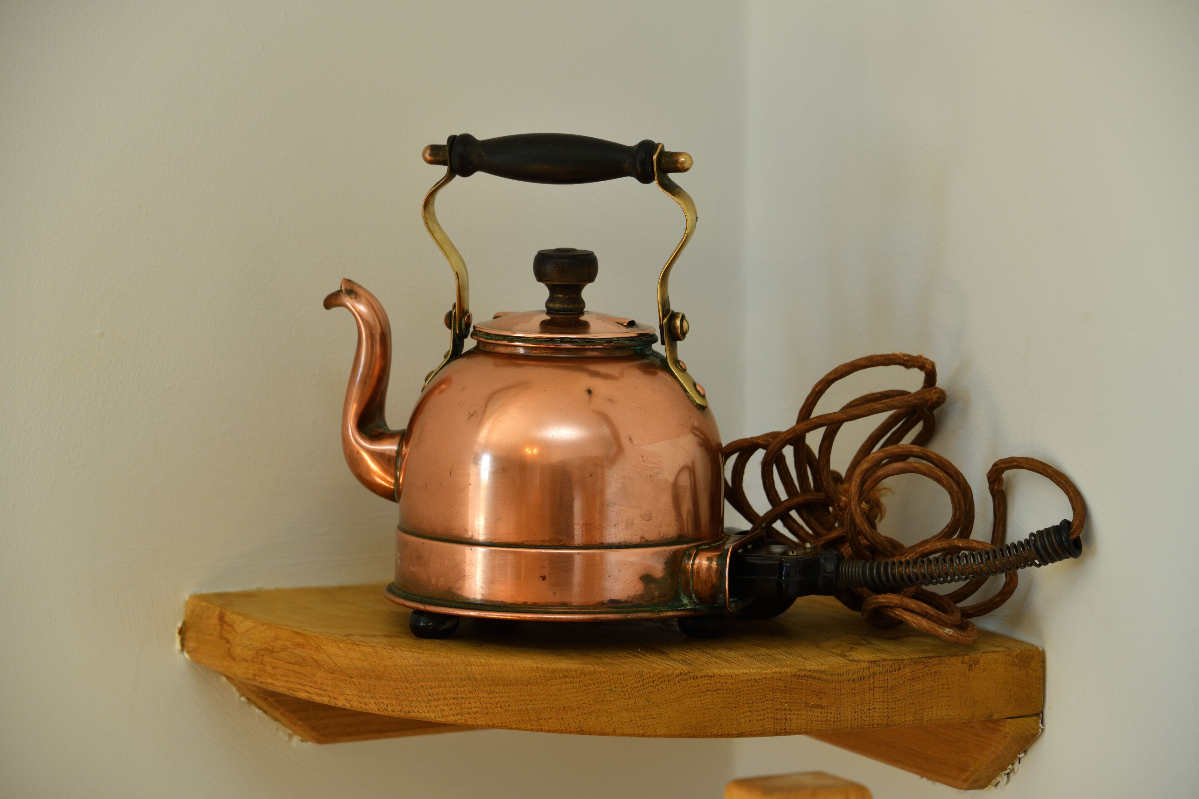 A copper kettle with a black handle and a black cord on a wooden shelf.