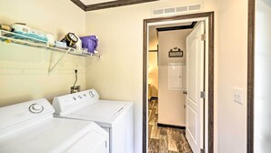 A laundry area with two white washers and dryers, a shelf with laundry supplies, and a hallway leading to a room with a wooden floor. - 'Pelican Place' Coastal Cottage Walk to Back Sound (Harkers Island)