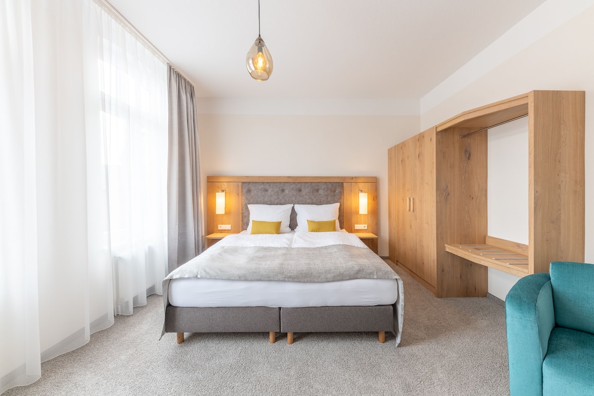 A neatly arranged hotel room with a large bed, bedside lamps, a wooden wardrobe, and a teal armchair.