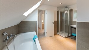 Un baño moderno con una bañera independiente, cabina de ducha con mampara de vidrio y un pasillo visible que conduce a otra habitación.