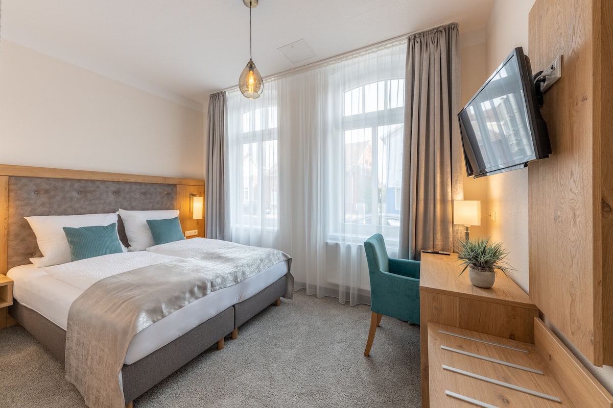 A hotel room with a bed, a desk with a chair, a television, and a window with curtains.
