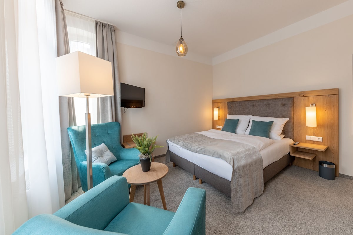 A hotel room with a bed, two armchairs, a small table with a plant, a TV on the wall, and a lamp.