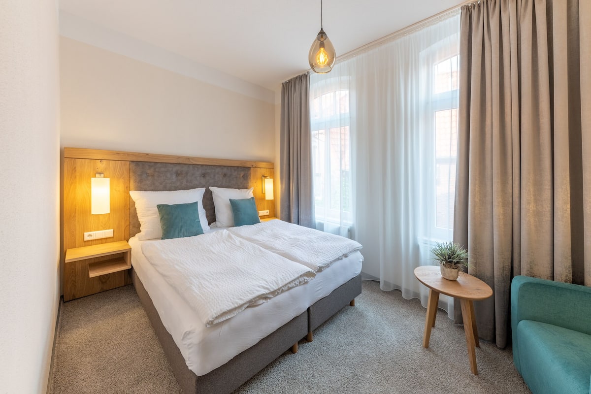 A hotel room with a large bed, bedside lamps, a small table with a plant, and a green armchair.