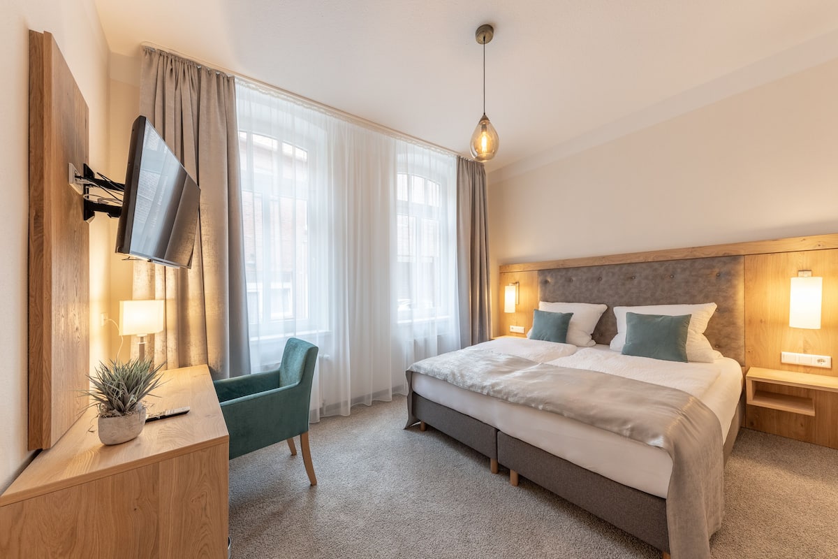 A modern hotel room with a large bed, a TV mounted on the wall, a desk with a lamp, and a potted plant.