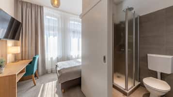 A modern hotel room with a bed, a desk with a chair, a TV, a shower, and a toilet.