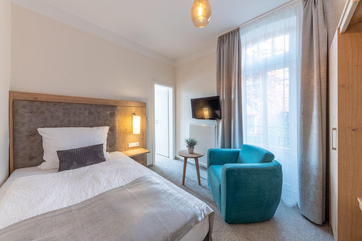 A hotel room with a large bed, a small table with a plant, a blue armchair, and a TV on the wall.