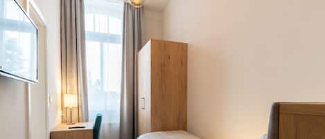 A compact hotel room with a single bed, a desk with a chair, a wooden wardrobe, a small trash bin, and a window with curtains.