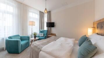 A hotel room with a bed, a teal armchair, a small table with a lamp, and a TV on the wall.