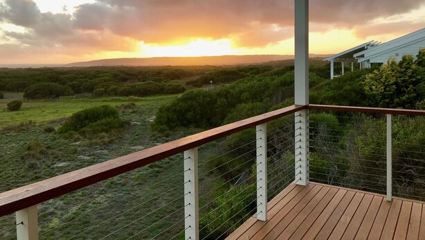A wooden deck with a railing overlooking a landscape with dense vegetation and a distant view of a building with a white roof. - Sea Salt Beach House | Luxury Coastal Escape for 12 (Goolwa Beach)