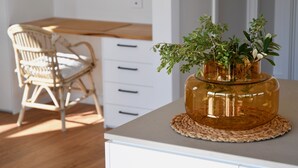 A room with a wooden dining table, a wicker chair, a white cabinet with drawers, and a glass vase with green plants on a woven placemat. - Sea Salt Beach House | Luxury Coastal Escape for 12 (Goolwa Beach)