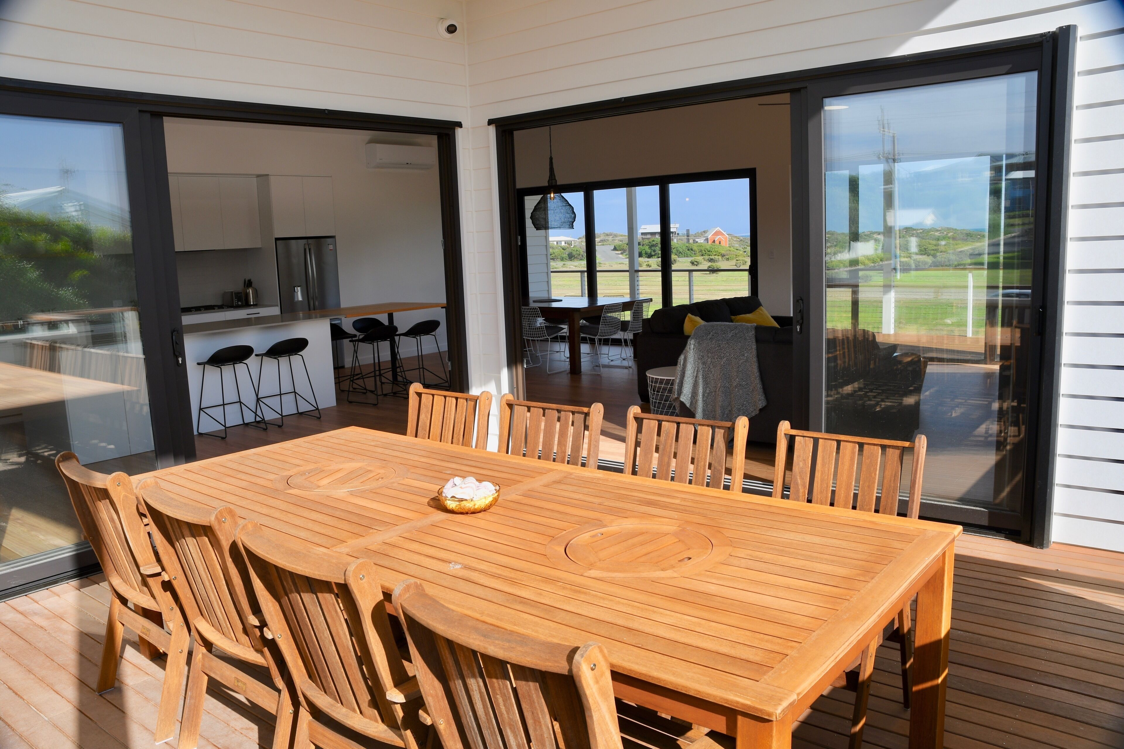 A wooden deck with a dining table and chairs, a kitchen area with modern appliances, and a large sliding glass door leading to a patio with a view of the countryside.