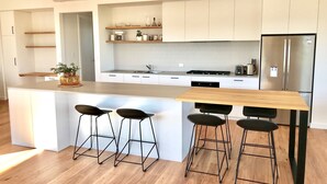 A modern kitchen with white cabinetry, a stainless steel refrigerator, a wooden island with bar stools, and a built-in oven. - Sea Salt Beach House | Luxury Coastal Escape for 12 (Goolwa Beach)