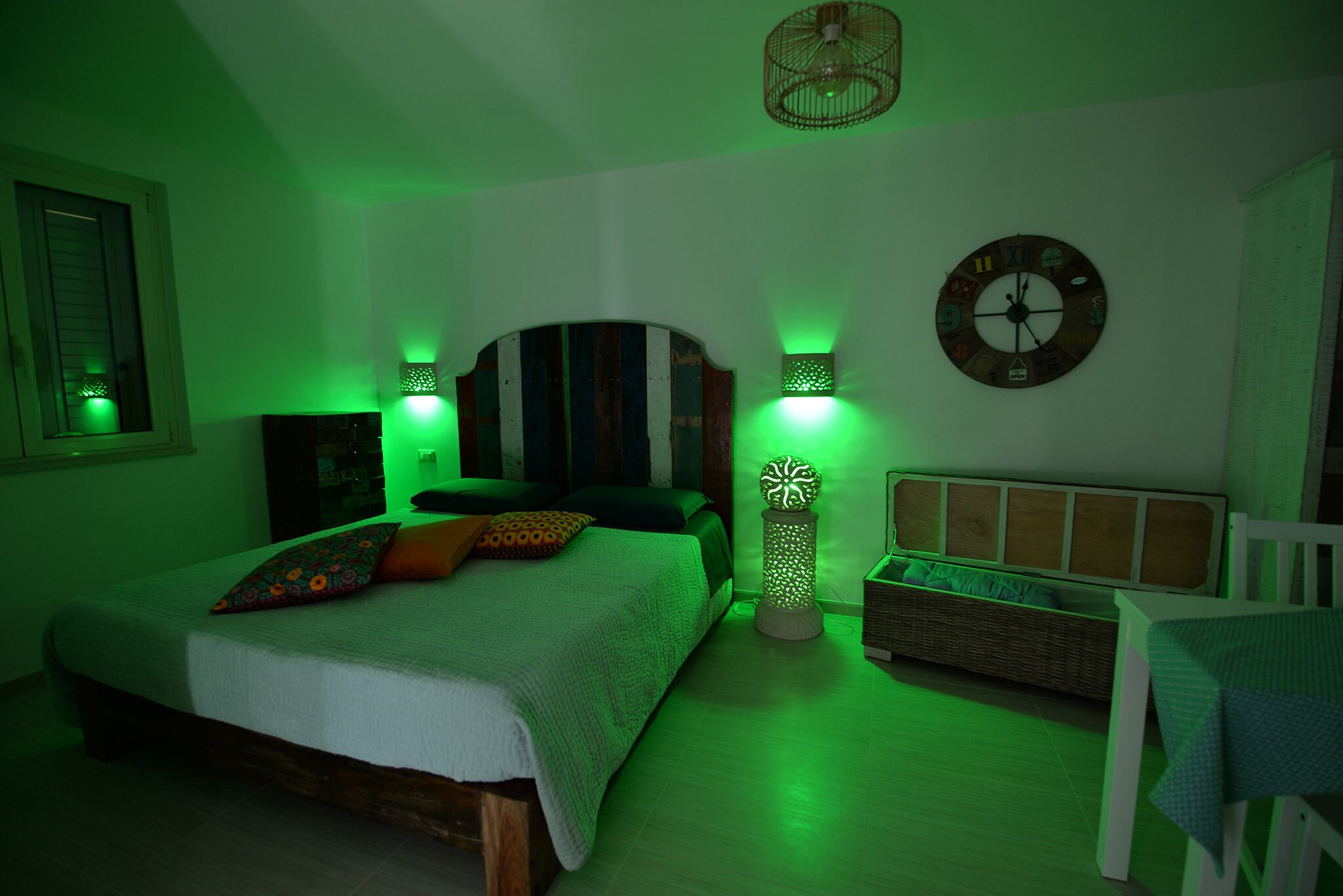 A bedroom with a bed, a nightstand, a wicker bench, a wall clock, and green ambient lighting.