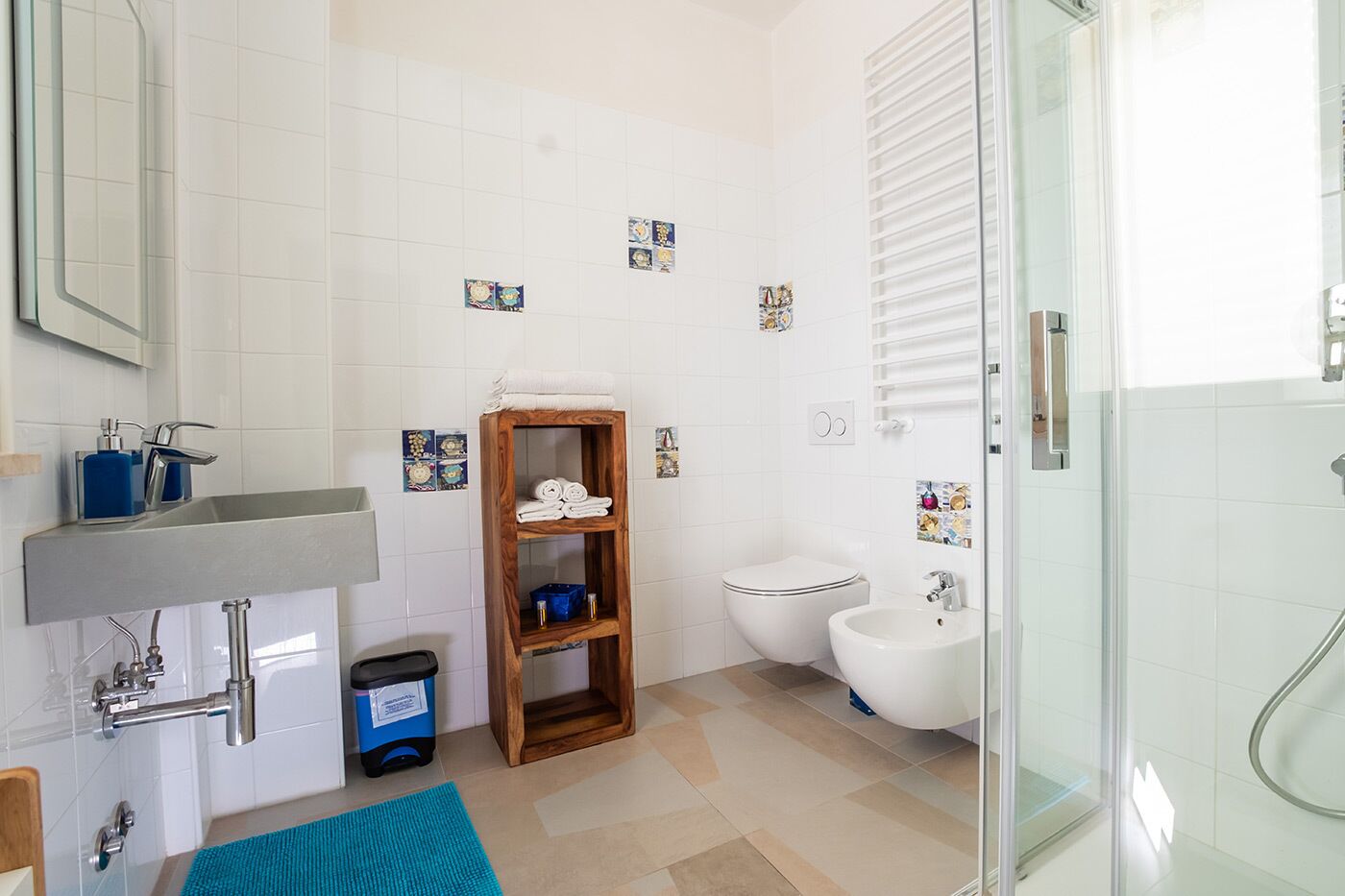 Een moderne badkamer met een wastafel, toilet, douche en bidet. Er is een houten plank met handdoeken, een prullenbak en decoratieve tegels op de muur.
