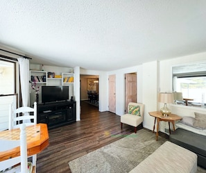 A living room with a TV, wooden furniture, a dining area, and a window with a view.