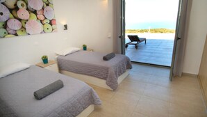 Une chambre d’hôtel avec deux lits, une vue sur la mer par une porte ouverte, une petite table avec une plante et une chaise à l’extérieur.