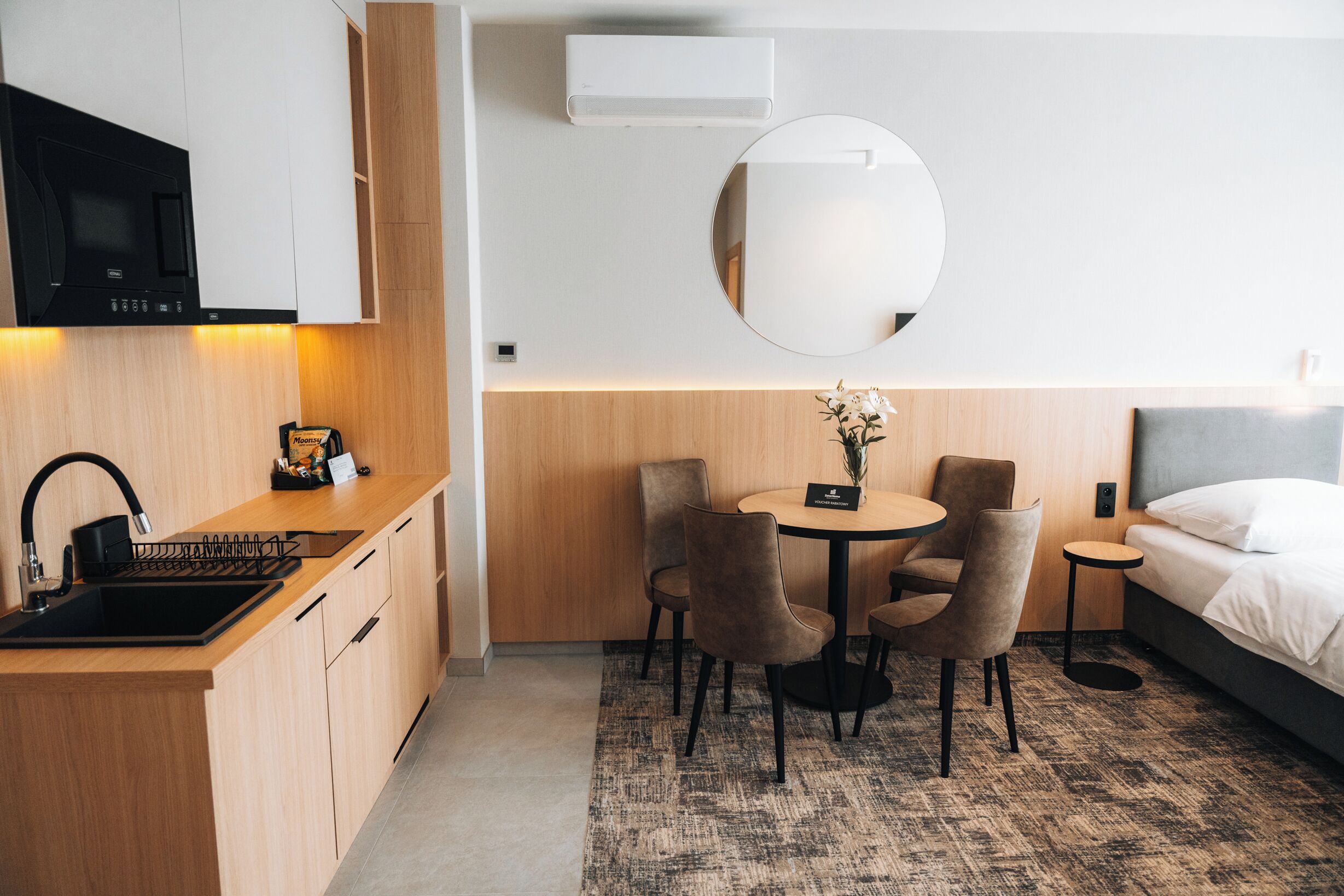 A modern hotel room with a kitchen area, a round dining table with chairs, a bed, and a round mirror on the wall.