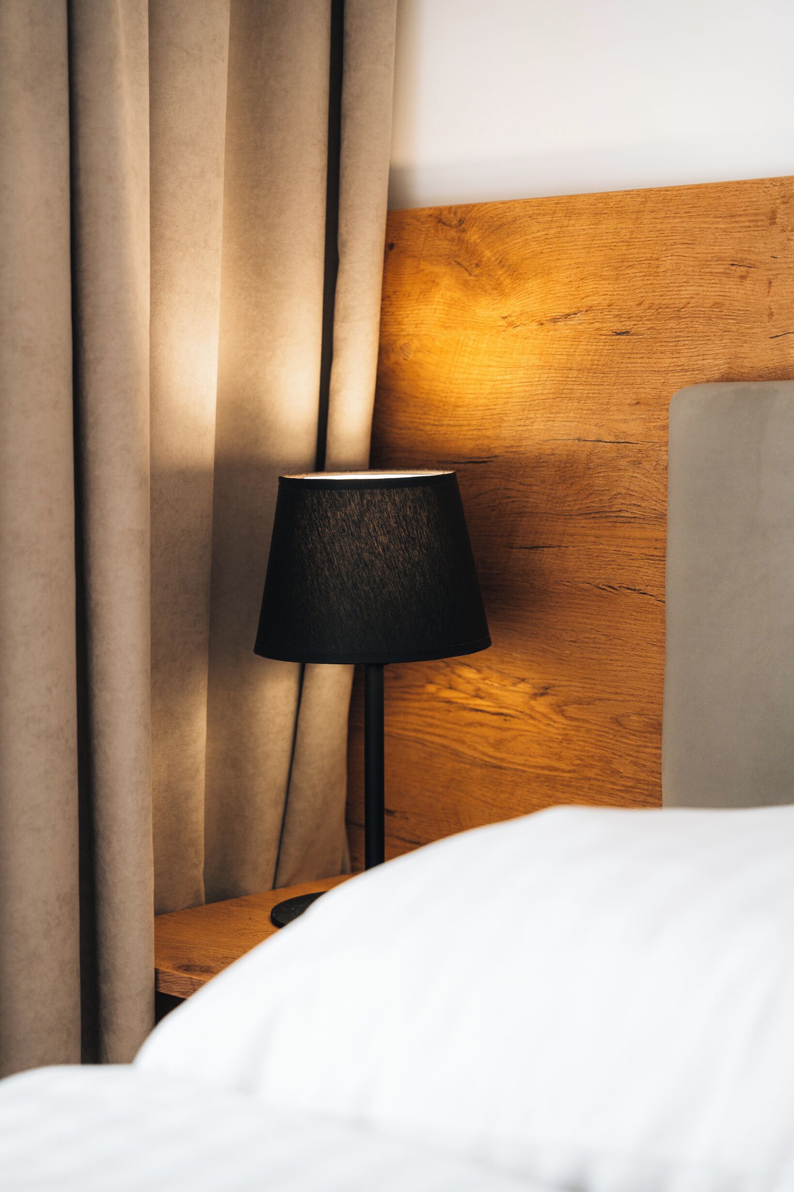Een bed met witte lakens, een houten nachtkastje, een zwarte lamp en beige gordijnen.