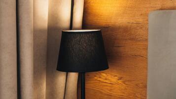 Een bed met witte lakens, een houten nachtkastje, een zwarte lamp en beige gordijnen.