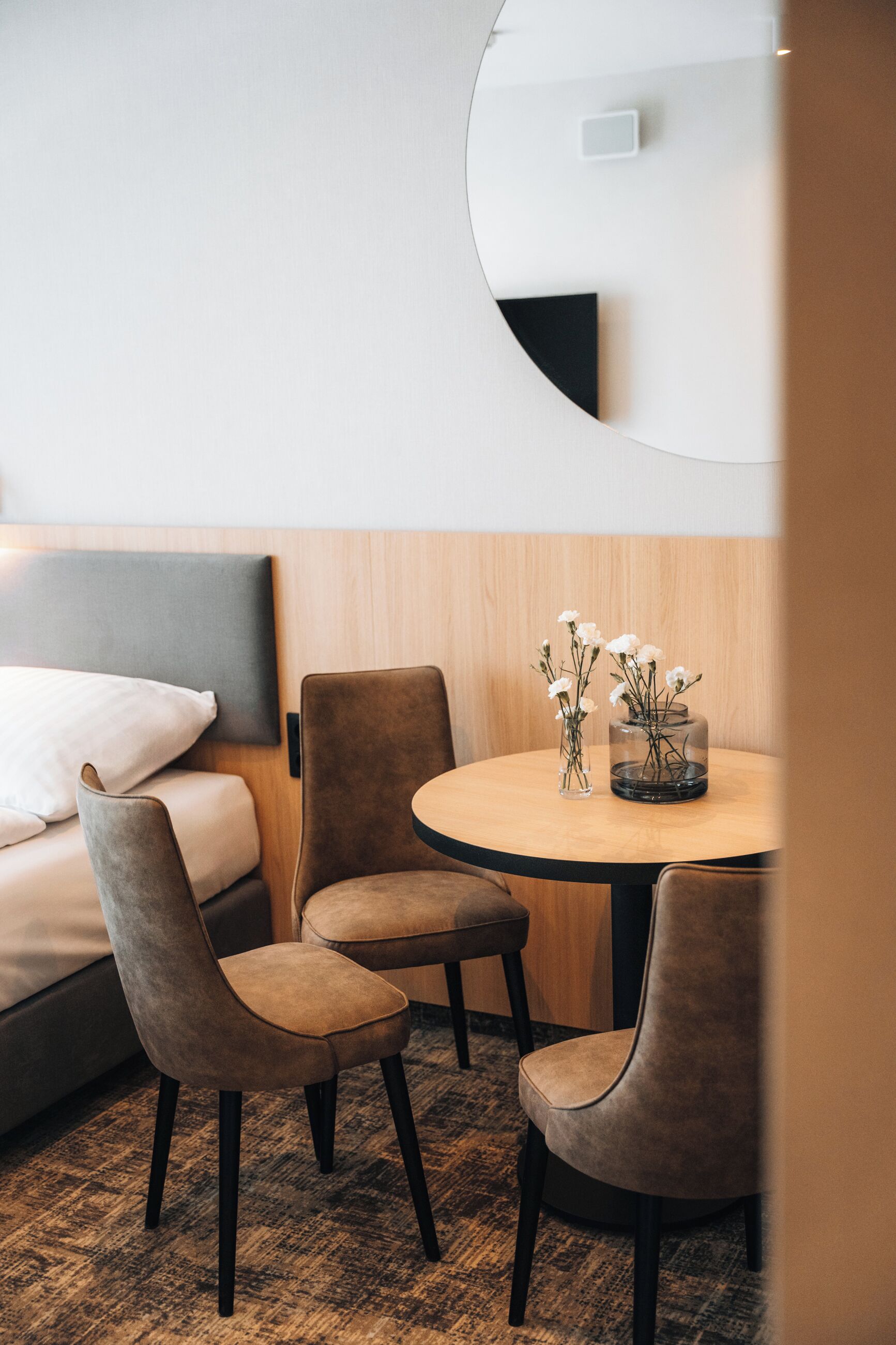 Ett modernt hotellrum med en säng, ett runt matbord med stolar och en vas med blommor.