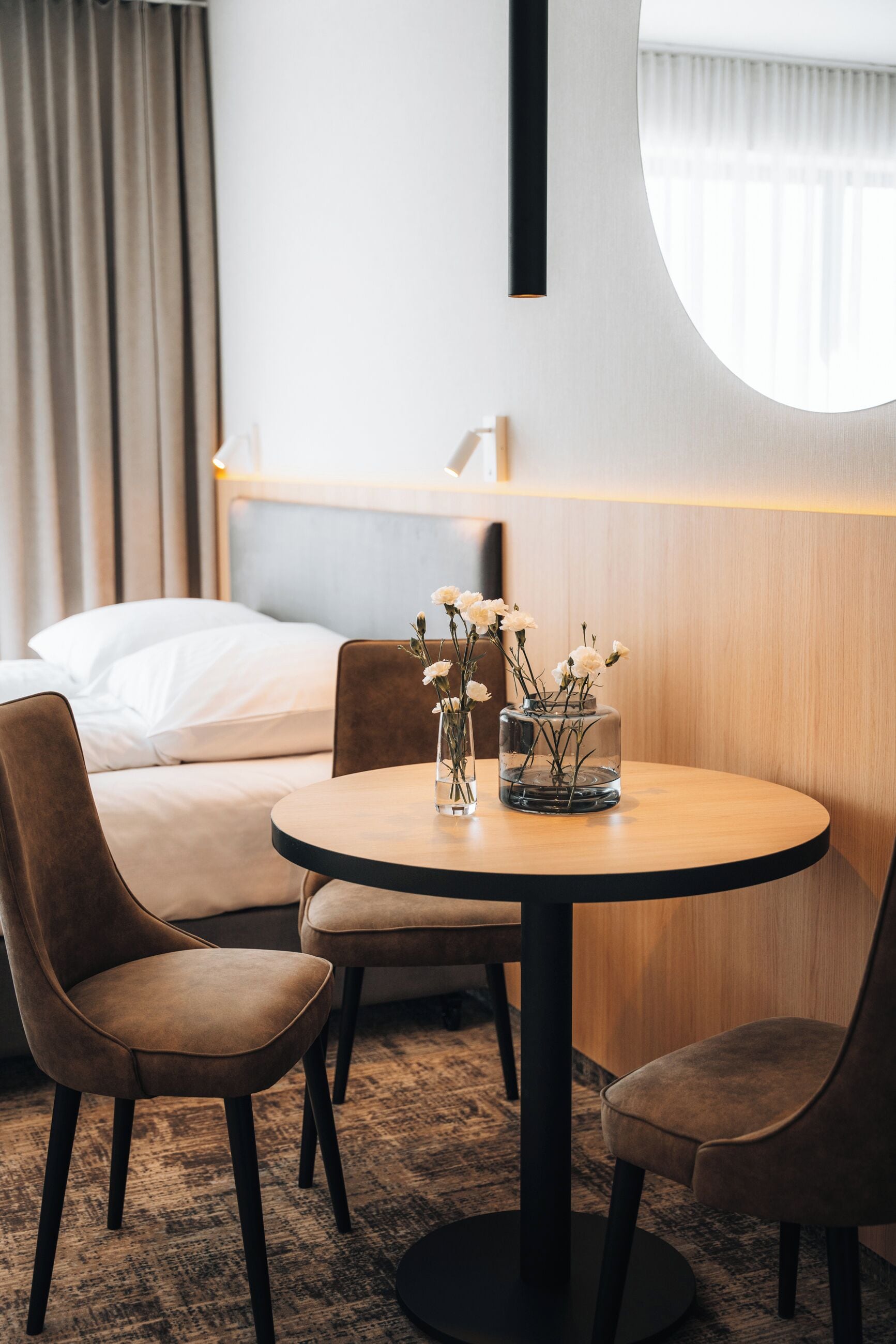 Ett hotellrum med ett runt träbord, två bruna stolar, en säng med vita sängkläder, en rund spegel och en vas med vita blommor.