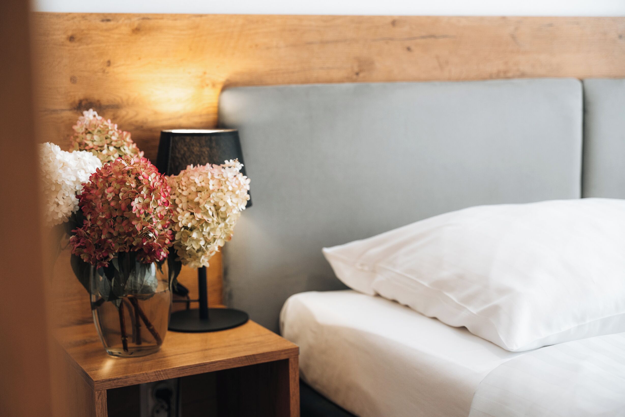 A neatly made bed with white linens, a bedside table with a lamp and a vase of flowers, and a wooden headboard.