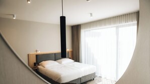 A modern hotel room with a curved mirror, a large bed, bedside tables, and a view of the city through large windows. - ZatorHome apartamenty (Zator)