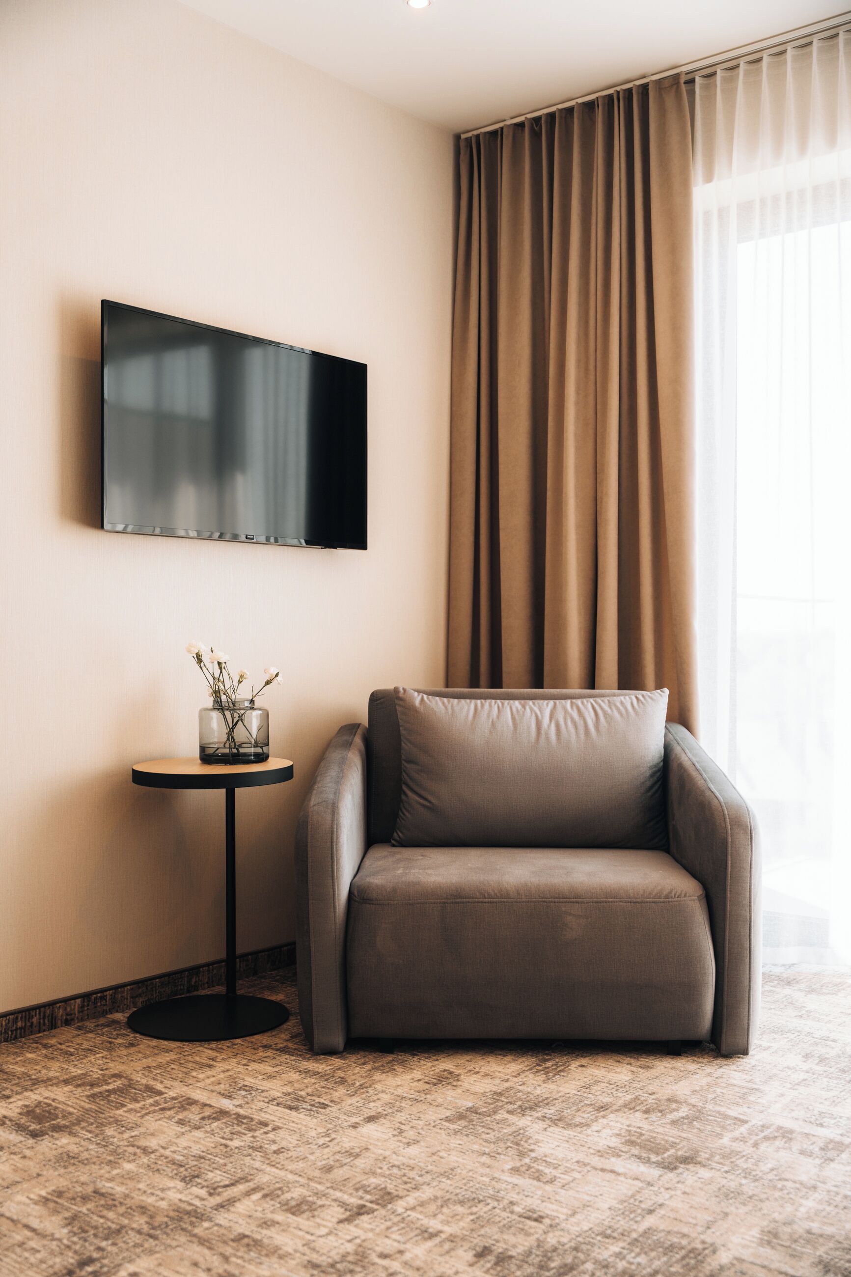 A modern hotel room with a flat-screen TV mounted on the wall, a round side table with a vase of flowers, a leather armchair, and a large window with curtains.