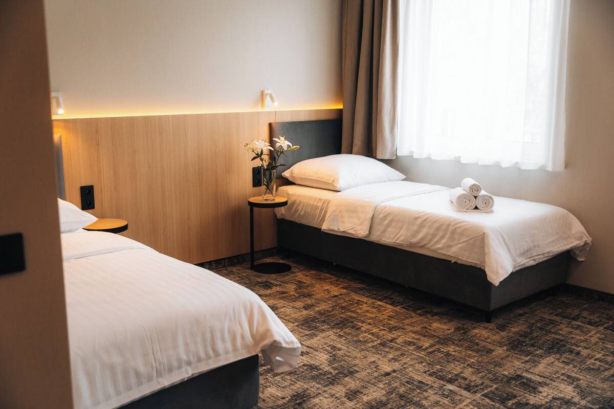 A hotel room with two beds, a bedside table with a lamp, a vase with flowers, and a window with curtains.