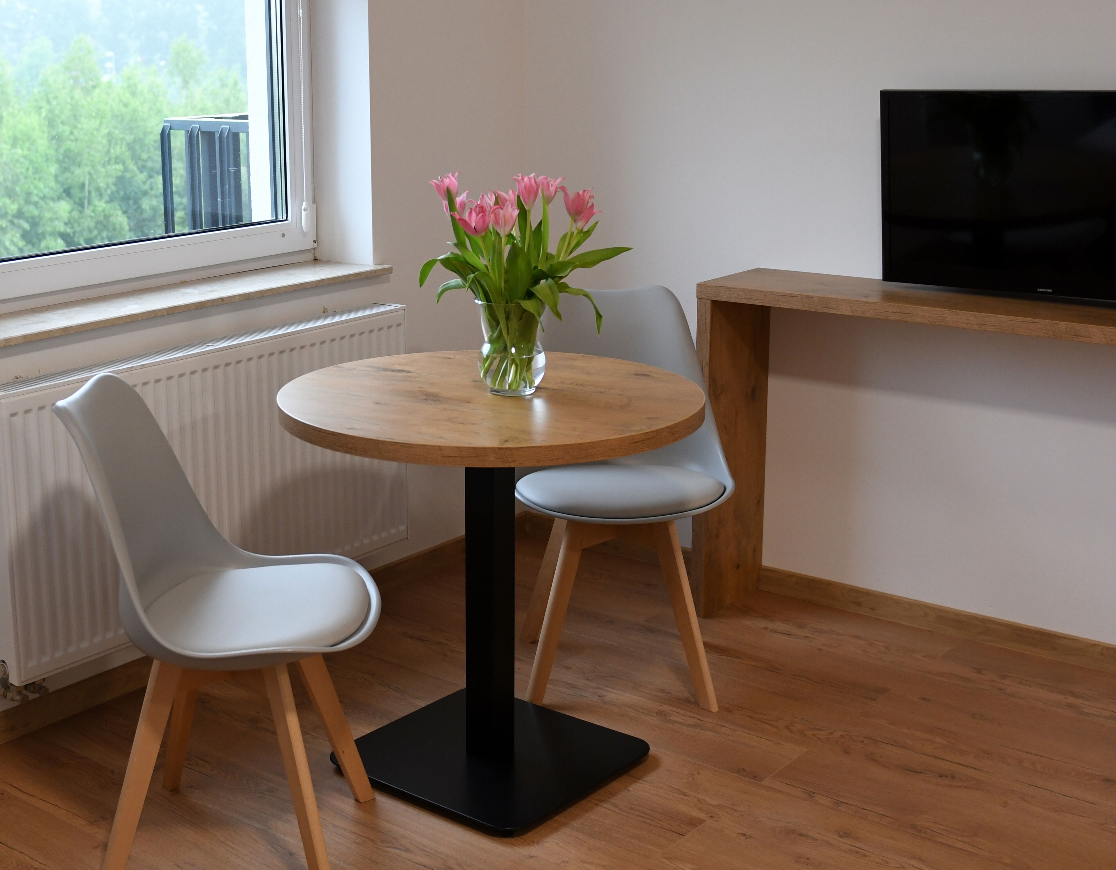 A small dining area with a round wooden table, two white chairs, and a vase of pink flowers. A television is mounted on the wall to the right.