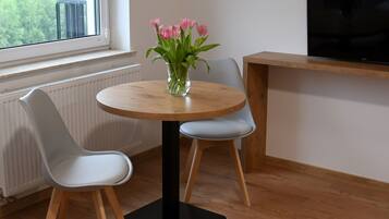 A small dining area with a round wooden table, two white chairs, and a vase of pink flowers. A television is mounted on the wall to the right.